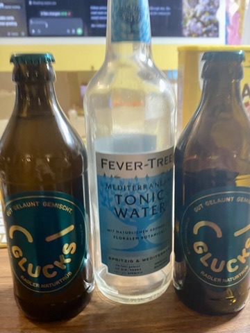 Three bottles standing together on a table: two brown Club-Mate bottles around a clear tonic-water bottle.