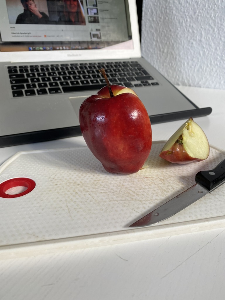 A bright red apple with a wedge cut away beside a kitchen knife, with an open laptop behind it.